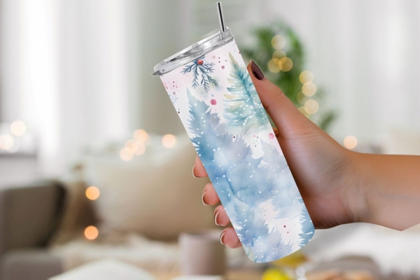 Hand hält Thermobecher mit winterlichem Design aus blauen Tannenbäumen und roten Akzenten, vor gemütlichem Lichterhintergrund.